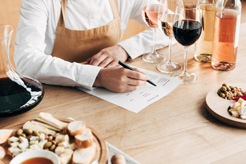 cropped view of sommelier in apron sitting at table and writing in wine tasting document