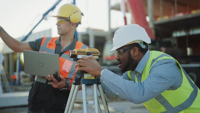 On the Commercial / Industrial Building Construction Site: Professional Engineer Surveyor Takes Measures with Theodolite, Worker Uses Laptop. In the Background Skyscraper Formwork Frames and Crane 