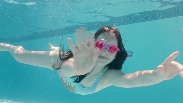 happy girl swimming underwater in pool smiling waving hand enjoying swim in crystal clear water wearing goggles enjoying summer 4k