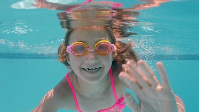 happy little girl swimming underwater in pool smiling waving hand enjoying swim in crystal clear water wearing goggles enjoying summer 4k
