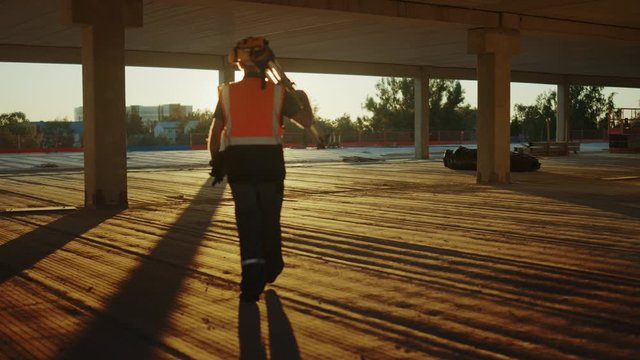 Inside of the Commercial / Industrial Building Construction Site: Engineer Surveyor Finishes Day's Job Carries Tripod Theodolite and Walks into Sunset