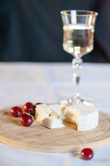Camembert from goat's milk cut into slices on a wooden board still life with sweet cherry berries. With a glass of white wine in the background
