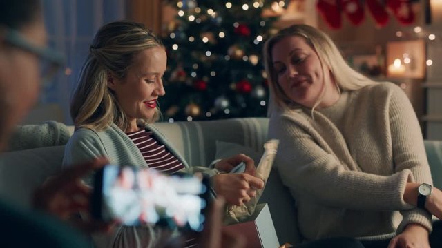 Christmas Family Opening Presents Pregnant Woman Unwrapping Gift Enjoying Surprise Baby Clothes Hugging Sister Celebrating Festive Holiday With Friend Taking Video Using Smartphone 4k