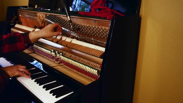 Technician tuning an upright piano by playing black and white keys and using lever and tools to tighten or loosen the strings to produce proper musical pitch.