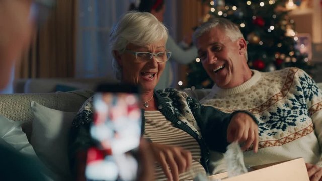 grandmother opening christmas gift with surprise sexy bra laughing enjoying funny joke with family taking video using smartphone celebrating festive holiday at home 4k