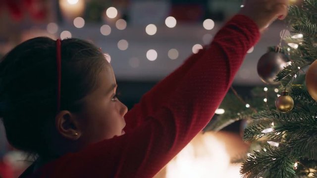 cute little girl decorating christmas tree with beautiful ornaments and baubles enjoying festive decorations at home on calm winter evening by fireplace 4k footage