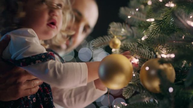 little girl with father decorating christmas tree with beautiful ornaments and baubles child helping dad hang festive decorations at home on calm evening 4k footage