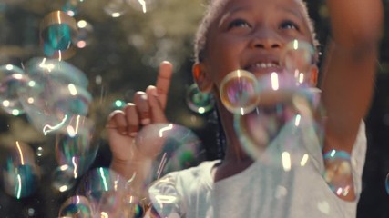 little african american girl playing with soap bubbles blowing playfully cute child having fun summer day playing bubble popping game in park 4k - Powered by Adobe
