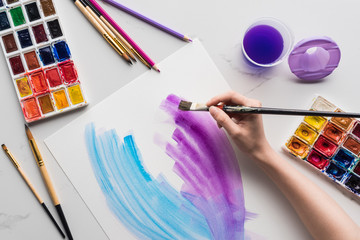 cropped view of artist drawing purple and blue watercolor brushstrokes on white paper on marble...