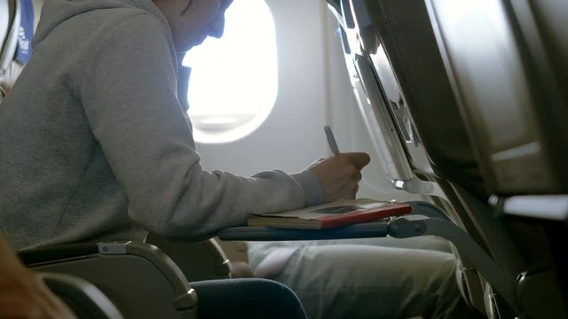 An attractive woman making notes in a notebook, sitting in a seat by the window on airplane. 4K