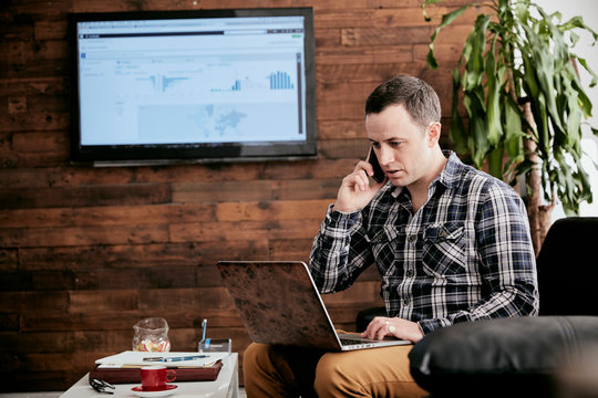 Businessman working on laptop while talking on smartphone in office