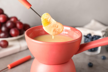 Dipping tangerine segment into pot with white chocolate fondue on table, closeup