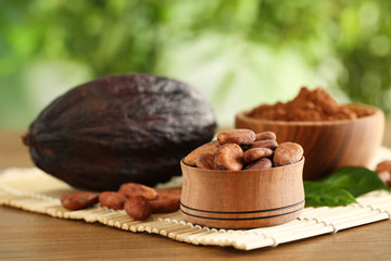 Wooden bowls of cocoa beans and powder near pod on table against blurred green background