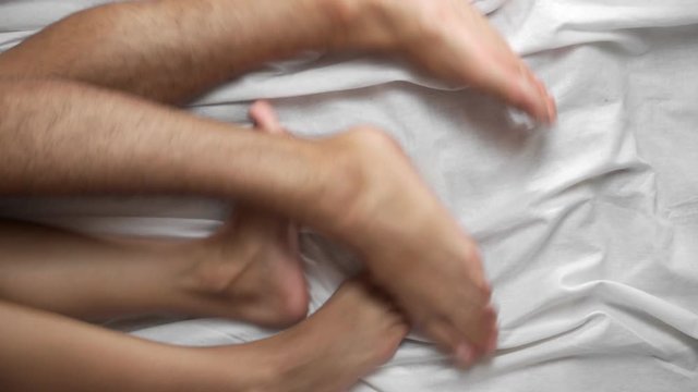 Top view. Female and male legs in bed on a white sheet are fighting among themselves. Close-up.