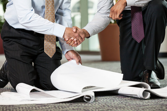 Low section of business people holding blueprint while shaking hands - Powered by Adobe