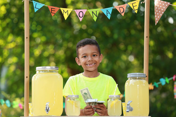 Cute little African-American boy with money at lemonade stand in park. Summer refreshing natural drink - obrazy, fototapety, plakaty