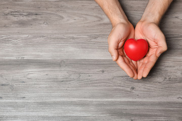 Young man holding red heart on grey wooden background, top view with space for text. Donation...