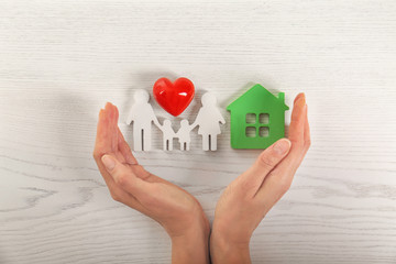 Woman holding hands near red heart and figures of house and family on white wooden background, top view