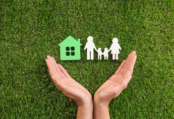 Woman holding hands near figures of house and family on green grass, top view