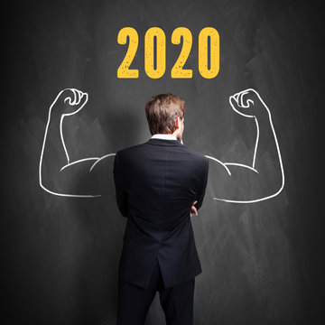 businessman standing in front of a blackboard with chalk message showing 2020 and drawn strong arms