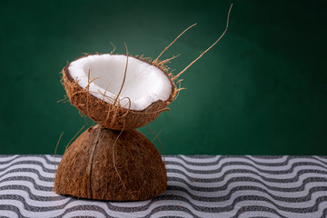 Coconut halves forming a chalice with the outer brown shell and inner white coco on a pattern surface against a dark green background with hairs sticking out. Studio concept of a natural cocktail cup.