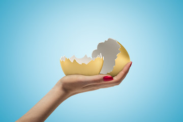Side closeup of woman's hand facing up and holding two parts of empty gold eggshell on light blue gradient background.
