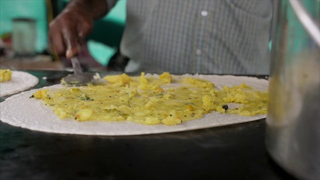 A road side food stall owner preparing masala dosa and adding mashed potatoes on the dosa batter . 