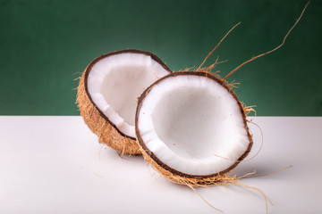 Two halves of a coconut with the brown outer shell and white coco interior on a white surface against a dark green background