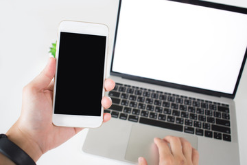 Laptops and hands are holding a smartphone with a blank screen on a white table inside the home or office. Isolated on white.