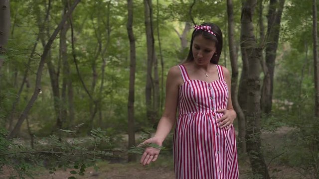 Pregnant girl in the park on a background of green trees. A girl with long dark hair in a striped white-red dress walks through the park.