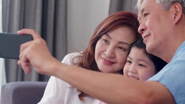 Asian grandparents selfie with granddaughter at home. Senior Chinese, grandpa and grandma happy spend family time relax using mobile phone with young girl kid lying on sofa in living room. Slow motion