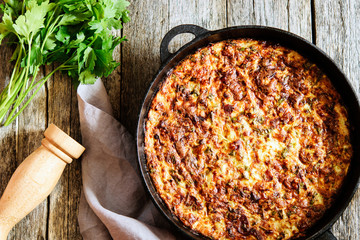 Zucchini Casserole with Bacon and Cheese in cast iron pan and parsley bunch and pepper mill on vintage wooden table. Bright sunlight. Selective focus