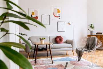 Minimal retro interior design of living room with grey couch, design armchair vintage coffee table, lamp and stylish decorations. Bright room, brwon wooden parquet and poster gallery on the wall.