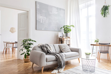 Stylish scandinavian living room with design furniture, plants, bamboo bookstand and wooden desk. Brown wooden parquet. Abstract painting on the white wall. Nice apartment. Modern decor of bright room - obrazy, fototapety, plakaty