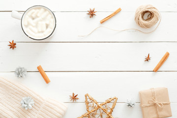 Fototapeta na wymiar Christmas minimal composition. Flatlay cup of hot chocolate with marshmallow, cinnamon sticks, Xmas decorations, gift box wrapped kraft paper, knitted hat on wooden white background. Hygge Christmas