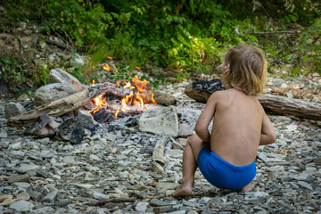 cute little boy in the forest sitting near the fire in the summer, view from the back