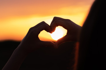 Silhouette of a young girl holding sun in her hands shaped like heart at sunset