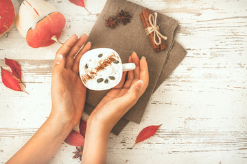 Woman hands hold a cup of spicy pumpkin latte, autumn hot drink white cup, with pumpkins, cinnamon and spices on old rustic wooden background copy space