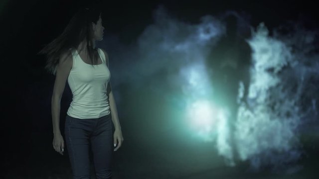 The female walking in a dark park against a man in a smoke behind. slow motion