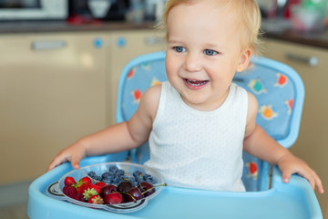 Adorable cute caucasian blond toddler boy enjoy tasting different seasonal fresh ripe organic berries sitting in highchair at home kitchen. Happy kid eating natural sweet healthy food as lunch snack