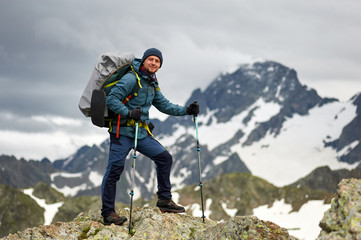 Hiker man stands on the cliff against the top of a mountain. Man has a backpack and trekking poles. Mountain trekking. Copy space
