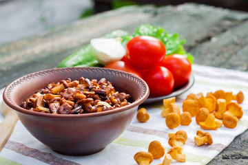 Fried chanterelles with onion in rustic bowl and plate with fresh vegetables for salad on background