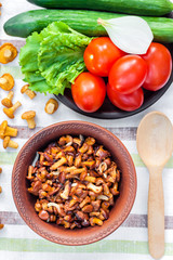 Fried chanterelles with onion in rustic bowl and plate with fresh vegetables for salad on background