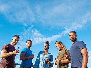 Happy friends standing with beer against blue sky. Group of young people relaxing after work. Celebration concept