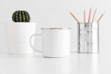 Enamel mug mockup with workspace accessories and a cactus in a pot on white table.