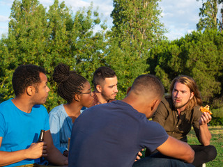 Side view of serious friends having picnic in park. Young people sitting on green grass and eating pizza. Concept of picnic