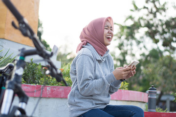 portrait of a hijab woman outdoors is taking a break holding a handphone and laughing