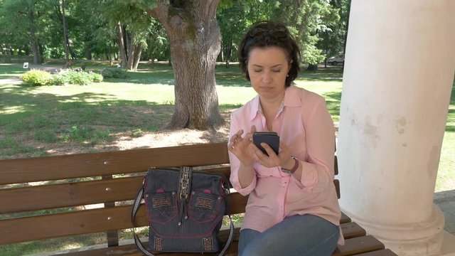 middle-aged brunette in a pink sweater and blue trousers sits in park on a bench and looks at the photo on her smartphone. On a sunny summer day blogger makes a post for social networks. camera motiom
