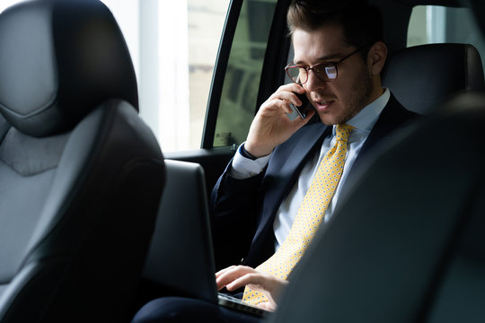 Young Businessman Sitting On Back Seat Of The Car, While His Chauffeur Is Driving Automobile.