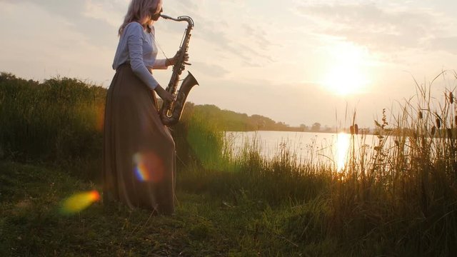 silhouette of a young woman playing on the saxophone near the riverbank at dawn, a beautiful girl in a long dress with a wind musical instrument in nature, a concept of hobby and relaxation